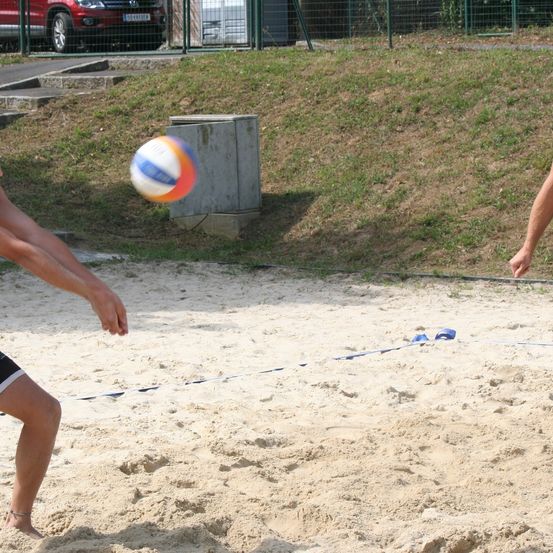Eine Person spielt Beachvolleyball auf sandigem Boden mit einem Netz vor sich. Der Ball ist in der Luft und ein Auto ist in der Ferne geparkt.