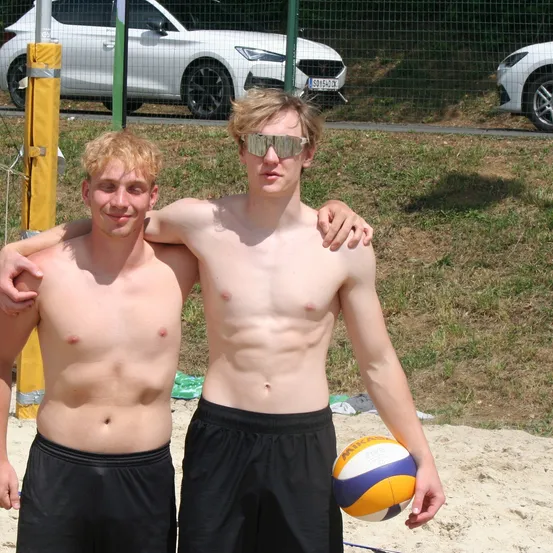 Zwei oberkörperlose junge Männer stehen auf dem Sand mit einem Volleyball in der Hand. Sie lächeln und posieren für ein Foto mit geparkten Autos hinter einem Zaun.