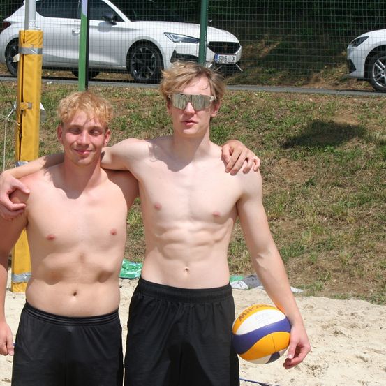 Zwei oberkörperlose junge Männer stehen auf dem Sand mit einem Volleyball in der Hand. Sie lächeln und posieren für ein Foto mit geparkten Autos hinter einem Zaun.