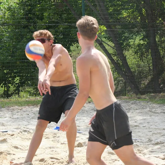 Zwei Jungen spielen Volleyball auf einem Sandplatz. Ein Junge bereitet sich darauf vor, den Ball zu schlagen, während der andere zusieht. Der Platz ist von einem Drahtzaun umgeben.