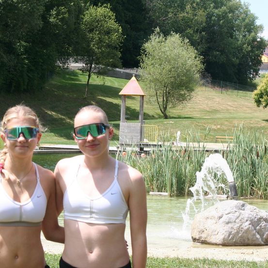 Zwei Mädchen in weißen Tanktops und Schwimmbrillen stehen neben einem Teich mit einer Fontäne. Bäume und Gras umgeben das Gebiet.