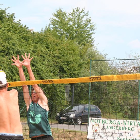 Zwei Männer spielen Volleyball in der Nähe einer Straße mit einem geparkten Auto. Ein Mann springt und erreicht das Netz. Bäume und ein Banner mit Text stehen dahinter.