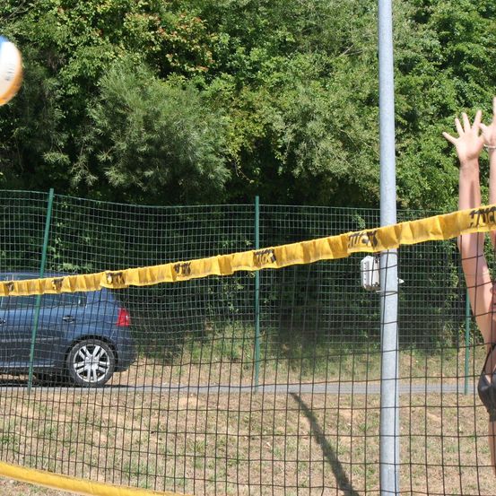 Eine Person spielt Volleyball mit einem Ball in der Luft in der Nähe eines Netzes, und ein Auto ist auf der Seite geparkt. Bäume sind im Hintergrund.