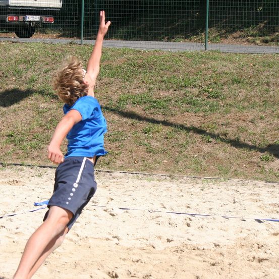Eine Person in einem blauen Shirt und Shorts spielt Volleyball in einem sandigen Bereich und springt mit erhobener rechter Hand. Dahinter steht ein Zaun, und ein Fahrzeug ist geparkt.