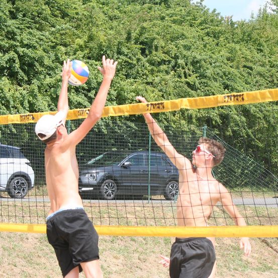 Zwei junge Männer spielen Volleyball auf einem Feld mit einem gelben Netz. Sie versuchen, einen Ball zu schlagen. Sie sind beide oberkörperfrei. Dahinter sind zwei Autos geparkt und ein buschiger Strauch.