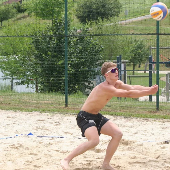 Ein Mann in Sonnenbrille und schwarzen Shorts spielt Volleyball auf einem Sandplatz. Der Ball ist in der Luft, und er bereitet sich darauf vor, ihn zu schlagen.