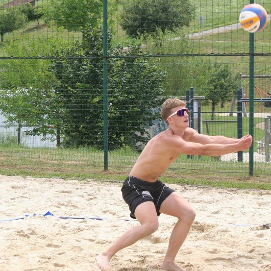 Ein Mann in Sonnenbrille und schwarzen Shorts spielt Volleyball auf einem Sandplatz. Der Ball ist in der Luft, und er bereitet sich darauf vor, ihn zu schlagen.