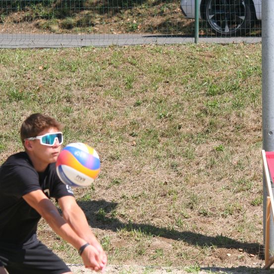 Ein Mann mit Sonnenbrille spielt Volleyball im Freien. Er versucht, einen bunten Ball zu treffen. Ein Auto und ein Zaun sind im Hintergrund zu sehen.