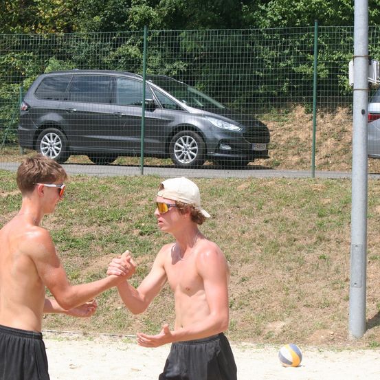Zwei junge Männer schütteln sich auf einem Strand die Hände, in der Nähe liegt ein Volleyball. Ein schwarzes Auto ist hinter einem Zaun geparkt.