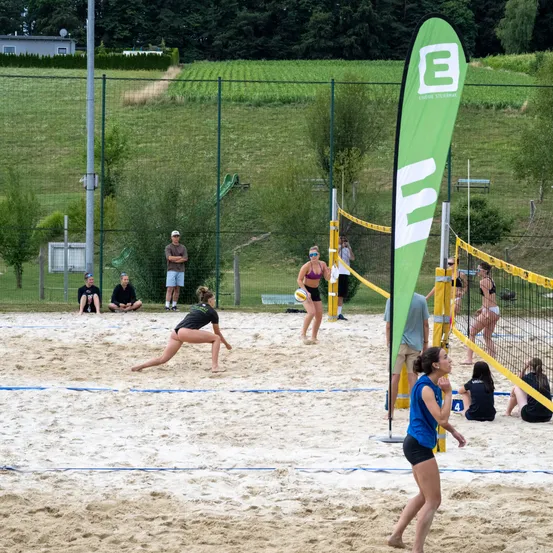 Eine Gruppe von Menschen spielt Volleyball auf einem sandigen Platz mit zwei Netzen, während andere von den Seitenlinien aus zuschauen.