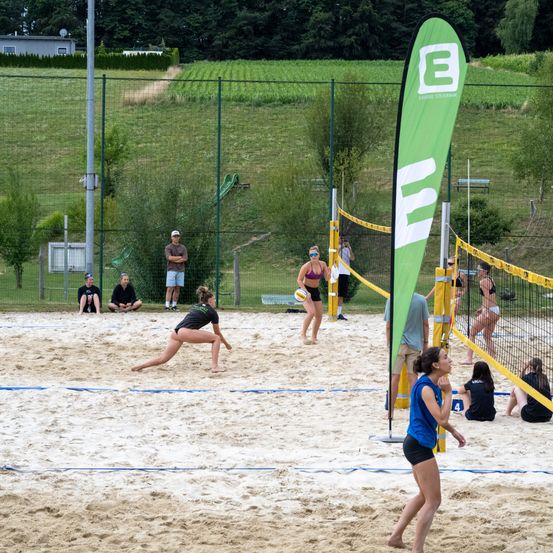 Eine Gruppe von Menschen spielt Volleyball auf einem sandigen Platz mit zwei Netzen, während andere von den Seitenlinien aus zuschauen.