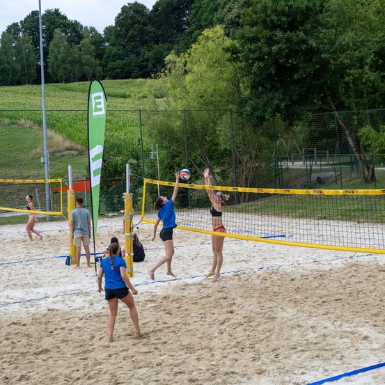 Eine Gruppe von Menschen spielt Volleyball auf einem sandigen Platz mit gelben Netzen. Sie tragen Sportbekleidung und sind in einem Spiel engagiert. Bäume und ein Zaun sind im Hintergrund.