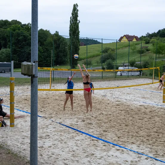 Zwei Frauen spielen Volleyball auf einem Sandplatz, eine in Rot und eine in Blau. Das Netz ist gelb, und eine weitere Spielerin sitzt im Sand. Bäume und ein Haus sind im Hintergrund.