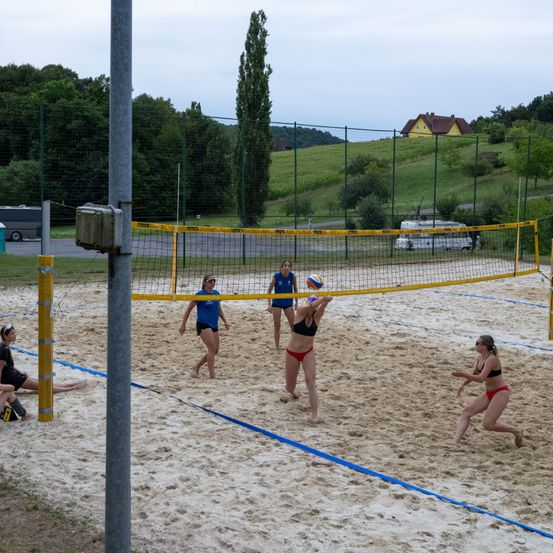 Eine Gruppe von Frauen spielt Beachvolleyball auf einem sandigen Spielfeld mit einem gelben Netz. Sie tragen Sportkleidung und befinden sich in verschiedenen Positionen, wahrscheinlich während eines Spiels. Im Hintergrund sind Bäume, ein Zaun und ein gelbes Haus in der Ferne zu sehen.