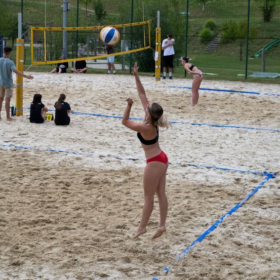 Ein Beachvolleyballspiel findet auf einem sandigen Platz statt. Eine Frau springt, um den Ball zu schlagen, während andere zuschauen und spielen.