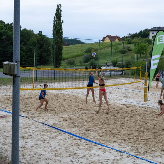 Eine Gruppe von Frauen spielt Beachvolleyball auf einem sandigen Platz mit einem gelben Netz. Eine Frau springt, um den Ball zu schlagen, während eine andere sich darauf vorbereitet, ihn zu empfangen. Bäume und Häuser sind im Hintergrund.