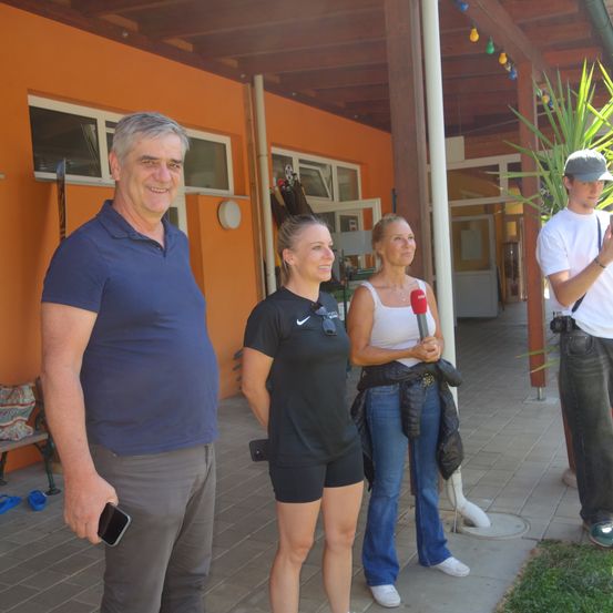 Drei Personen stehen auf einer Veranda. Ein Mann in einem blauen Shirt, eine Frau in Schwarz mit einem Mikrofon und ein Mann in Weiß mit einer Kamera. Sie scheinen zu unterhalten.