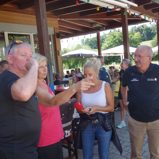 Ein Outdoor-Restaurant mit Menschen, einer mit Mikrofon, und eine Frau mit blonden Haaren spricht. Es gibt Bäume und Zelte im Hintergrund.