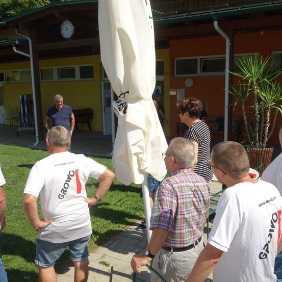 Mehrere Erwachsene in Freizeitkleidung versammeln sich vor einem Gebäude. Ein Mann steht in der Nähe einer Uhr, und andere schauen eine Pflanze in einem Topf an. Ein geschlossener Regenschirm ist sichtbar.