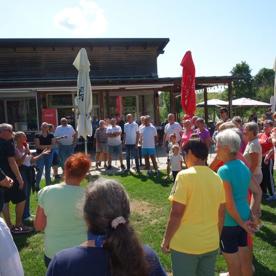 Eine Gruppe von Menschen versammelt sich vor einem Gebäude mit einem Coca-Cola-Schild. Einige stehen, andere sitzen und scheinen zu sprechen. Ein Schirm ist in der Nähe.