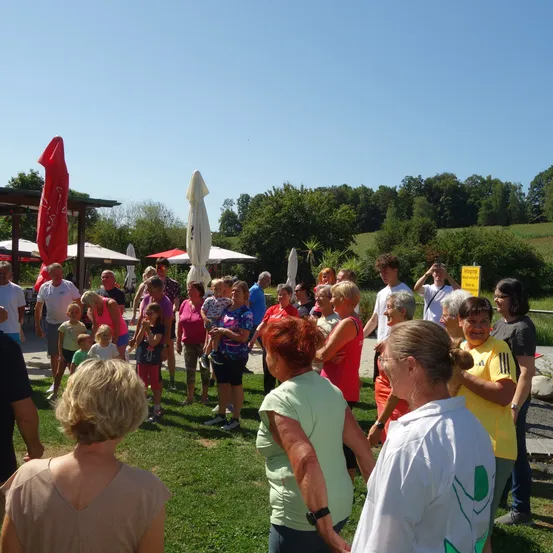 Eine Gruppe von Menschen versammelt sich in einem Außenbereich, wahrscheinlich einem Park. Sie unterhalten sich und einige halten sich an den Händen. Im Hintergrund befinden sich Bäume und Pflanzen.