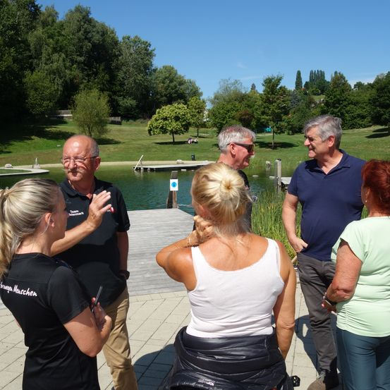 Eine Gruppe von Menschen steht in der Nähe eines Sees, eine Frau justiert ihr Haar. Zwei Männer tragen Sonnenbrillen und schwarze Hemden, während ein anderer Mann in einem blauen Hemd lächelt. Der See ist von Gras und Bäumen umgeben.