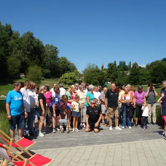 Eine Gruppe von Menschen steht draußen in der Nähe eines Teichs, einige kniend oder auf einer Holzplattform sitzend. Sie sind lässig gekleidet und scheinen für ein Foto zu posieren. Bäume und ein blauer Himmel sind im Hintergrund zu sehen.
