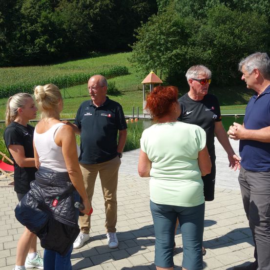 Eine Gruppe von Menschen steht in einem Kreis auf einem gepflasterten Weg. Sie unterhalten sich miteinander. Die Umgebung umfasst einen See, ein Pavillon und ein grasbewachsenes Feld.
