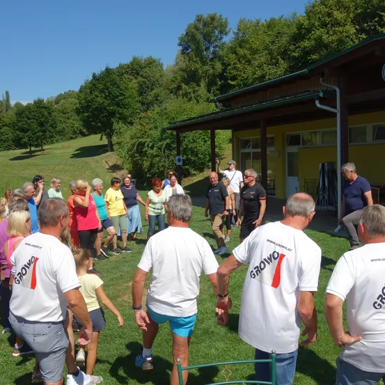 Eine Gruppe von Menschen in gleichen weißen T-Shirts versammelt sich auf einem Rasenfeld vor einem gelben Gebäude. Die Erwachsenen stehen, während ein Kind in der Nähe geht. Das Gebiet ist von Bäumen und Gras umgeben.