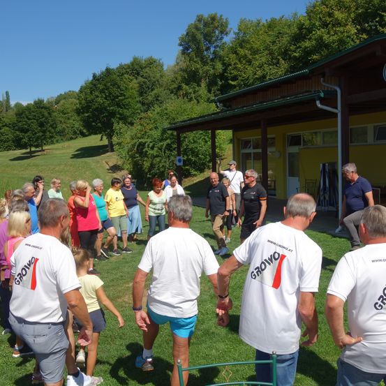 Eine Gruppe von Menschen in gleichen weißen T-Shirts versammelt sich auf einem Rasenfeld vor einem gelben Gebäude. Die Erwachsenen stehen, während ein Kind in der Nähe geht. Das Gebiet ist von Bäumen und Gras umgeben.