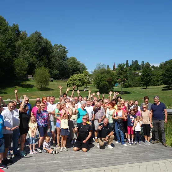 Eine Gruppe von Menschen posiert für ein Foto auf einer Holzterrasse mit grünem Hintergrund. Sie alle lächeln und winken mit den Händen. Einige knien, andere stehen. Sie tragen Freizeitkleidung. In der Ferne befinden sich Bäume und ein Turm.