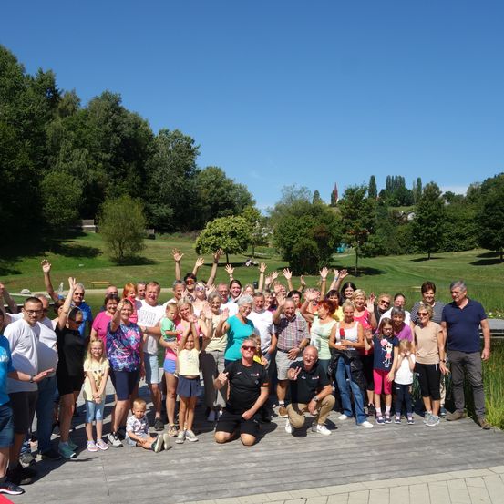 Eine Gruppe von Menschen posiert für ein Foto auf einer Holzterrasse mit grünem Hintergrund. Sie alle lächeln und winken mit den Händen. Einige knien, andere stehen. Sie tragen Freizeitkleidung. In der Ferne befinden sich Bäume und ein Turm.