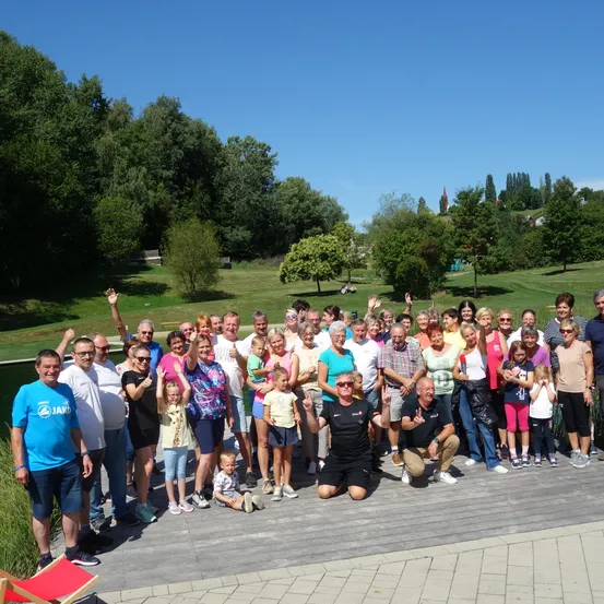 Eine große Gruppe von Menschen, einschließlich Erwachsener und Kindern, posiert für ein Foto auf einer Holzterrasse. Sie stehen und sitzen auf der Terrasse, einige winken. Der Hintergrund zeigt einen Rasenbereich mit Bäumen und einem Gewässer.