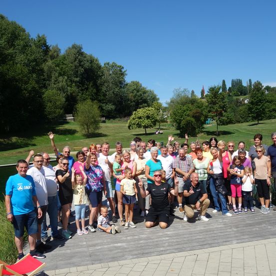 Eine große Gruppe von Menschen, einschließlich Erwachsener und Kindern, posiert für ein Foto auf einer Holzterrasse. Sie stehen und sitzen auf der Terrasse, einige winken. Der Hintergrund zeigt einen Rasenbereich mit Bäumen und einem Gewässer.