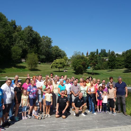 Eine große Gruppe von Menschen, überwiegend ältere Personen, posiert für ein Foto in einem Park an einem sonnigen Tag. Sie stehen auf einer Holzplattform mit einem Rasenfeld und Bäumen im Hintergrund.