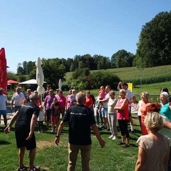 Eine Gruppe von Menschen in Freizeitkleidung, einige in roten Shirts, versammelt sich auf einem Rasenfeld mit Bäumen und einem entfernten Feld im Hintergrund.