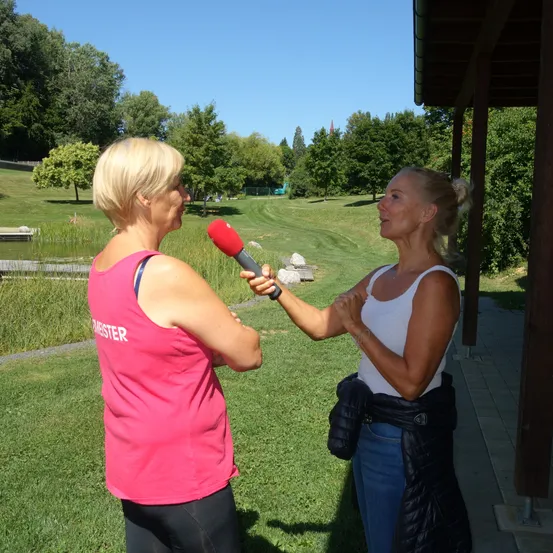 Zwei Frauen sind draußen in einem Park, eine hält ein Mikrofon und interviewt die andere. Hinter ihnen ist ein Rasenbereich, ein Teich und Bäume. Der Himmel ist klar.