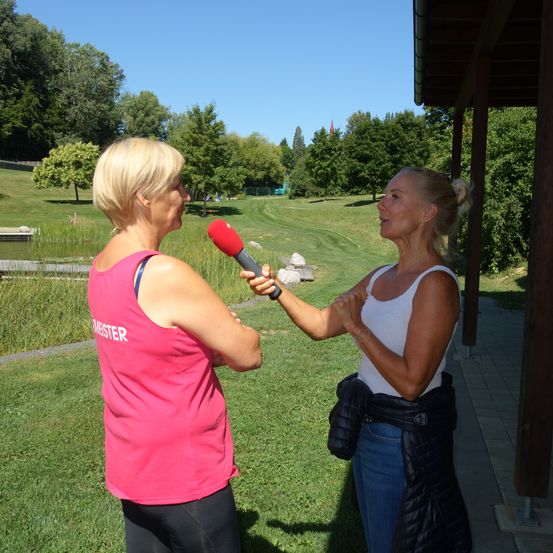 Zwei Frauen sind draußen in einem Park, eine hält ein Mikrofon und interviewt die andere. Hinter ihnen ist ein Rasenbereich, ein Teich und Bäume. Der Himmel ist klar.