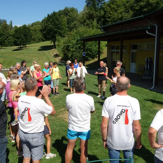 Eine Gruppe von Menschen in weißen T-Shirts mit dem Wort Grovo stehen auf einem grünen Rasen und klatschen in die Hände. Vor ihnen steht ein Gebäude mit einem grünen Dach.