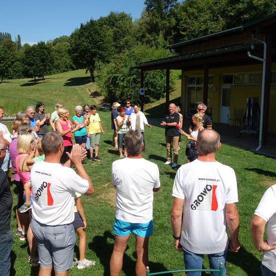 Eine Gruppe von Menschen in weißen T-Shirts mit dem Wort Grovo stehen auf einem grünen Rasen und klatschen in die Hände. Vor ihnen steht ein Gebäude mit einem grünen Dach.