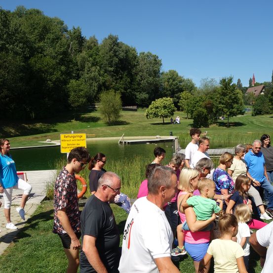 Eine Gruppe von Menschen versammelt sich um einen Teich in einem Park. Einige sitzen auf dem Gras, während andere in der Nähe des Wassers stehen. Ein gelbes Schild warnt vor Schwimmbeschränkungen.