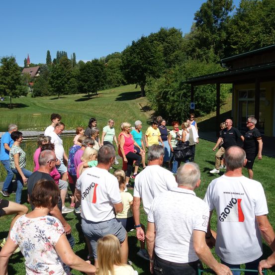Eine Gruppe von Menschen auf einer Rasenfläche, einige tragen weiße T-Shirts mit einem roten Logo, nehmen an einer Outdoor-Veranstaltung bei klarem Himmel mit Bäumen und einem Gebäude im Hintergrund teil.