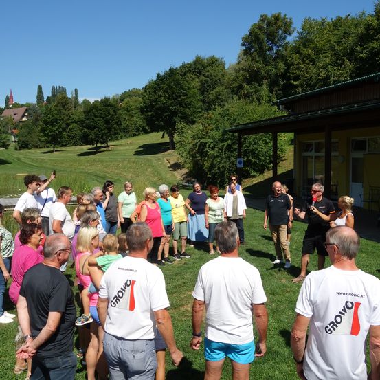 Eine Gruppe von Menschen versammelt sich auf einem Rasen, möglicherweise für eine Veranstaltung. Einige tragen Turnschuhe und Brillen, und einige halten Kameras. Sie stehen vor einem Gebäude mit gelber Wand.