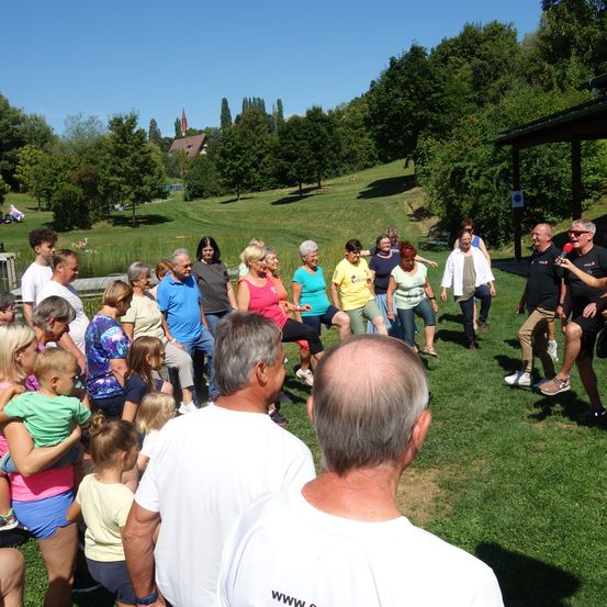 Eine Gruppe von Menschen versammelt sich auf einem Rasenfeld, einige tanzen und andere schauen zu. Sie sind lässig gekleidet. Der Hintergrund zeigt einen Hügel mit Bäumen und einem Haus.