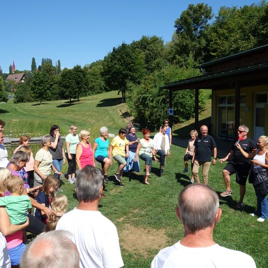 Eine Gruppe von Menschen, einige tanzen, auf einer Rasenfläche mit einem gelben Gebäude, umgeben von Bäumen und einem Hügel.