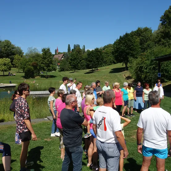 Eine Gruppe von Menschen steht in einem grasbewachsenen Parkbereich mit einem Teich und Bäumen im Hintergrund. Sie sind lässig gekleidet und einige halten Kinder.