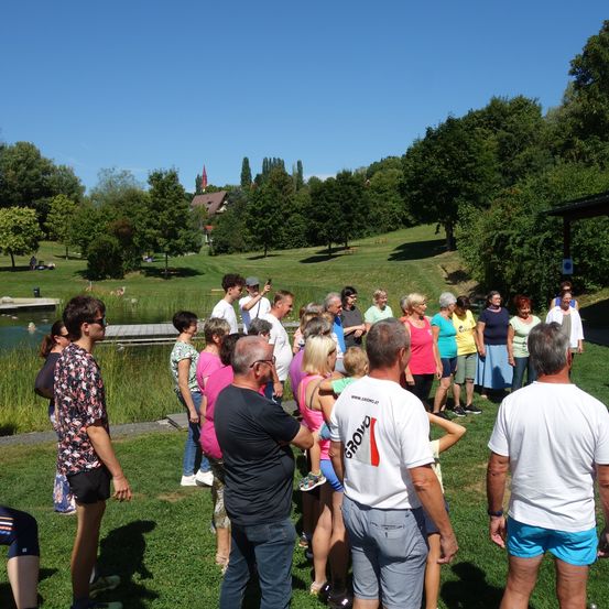 Eine Gruppe von Menschen steht in einem grasbewachsenen Parkbereich mit einem Teich und Bäumen im Hintergrund. Sie sind lässig gekleidet und einige halten Kinder.