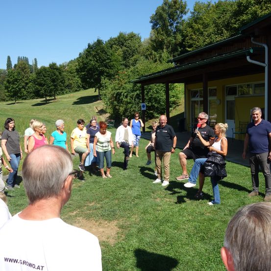 Eine Gruppe von Menschen versammelt sich im Freien in einem Park. Einige stehen, andere sitzen. Ein Mann hält ein Mikrofon und hält möglicherweise eine Rede. Bäume und ein Gebäude sind im Hintergrund zu sehen.