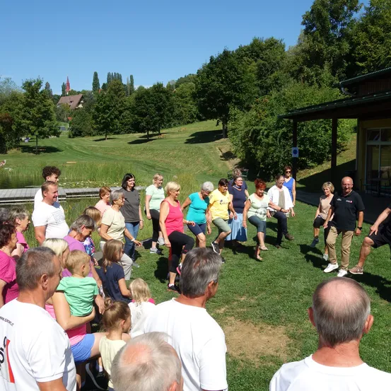 Eine Gruppe von Menschen versammelt sich auf einer Rasenfläche, einige gehen und andere stehen. Im Hintergrund befindet sich ein Gebäude mit Giebeldach. Die Szene spielt sich in einem Park mit Bäumen und einem klaren blauen Himmel ab.