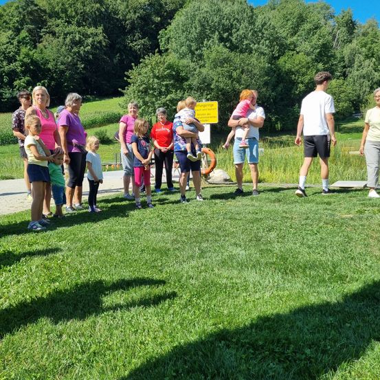 Eine Gruppe von Erwachsenen und Kindern steht auf einem Grasfeld. Einige halten Kinder, und ein Rettungsring befindet sich in der Nähe. Bäume und Büsche sind im Hintergrund.